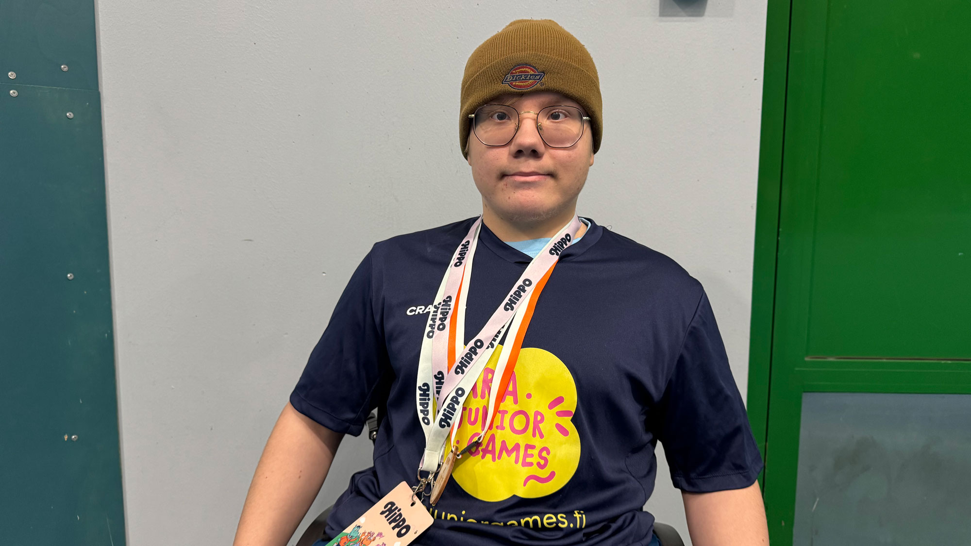 Pipopäinen pyörätuolia käyttävä poika Para Junior Games -kisapaidassa ja silmälaseissa hymyilee kameralle kisapaikalla lähikuvassa.